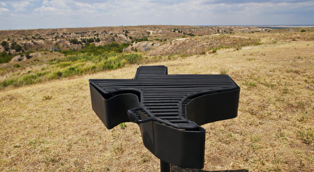 Una parrilla de barbacoa con la forma de Texas, con el paisaje agreste de Texas en el fondo